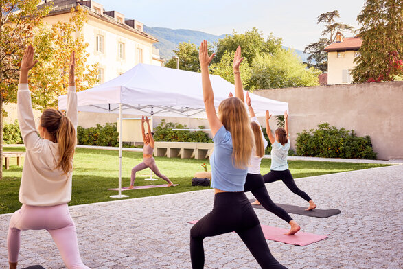 The white folding gazebo is in use at a yoga class. The yoga instructor is standing under it. In front of the gazebo are the participants in the yoga class.