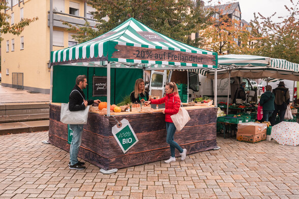 The market tent is fully printed and is used at the weekly market and farmers' market. 2 customers look at the products.