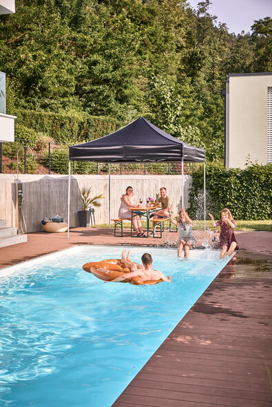 The black folding gazebo stands by the pool in the garden. Friends sit under it in the shade and have a pool and garden party.